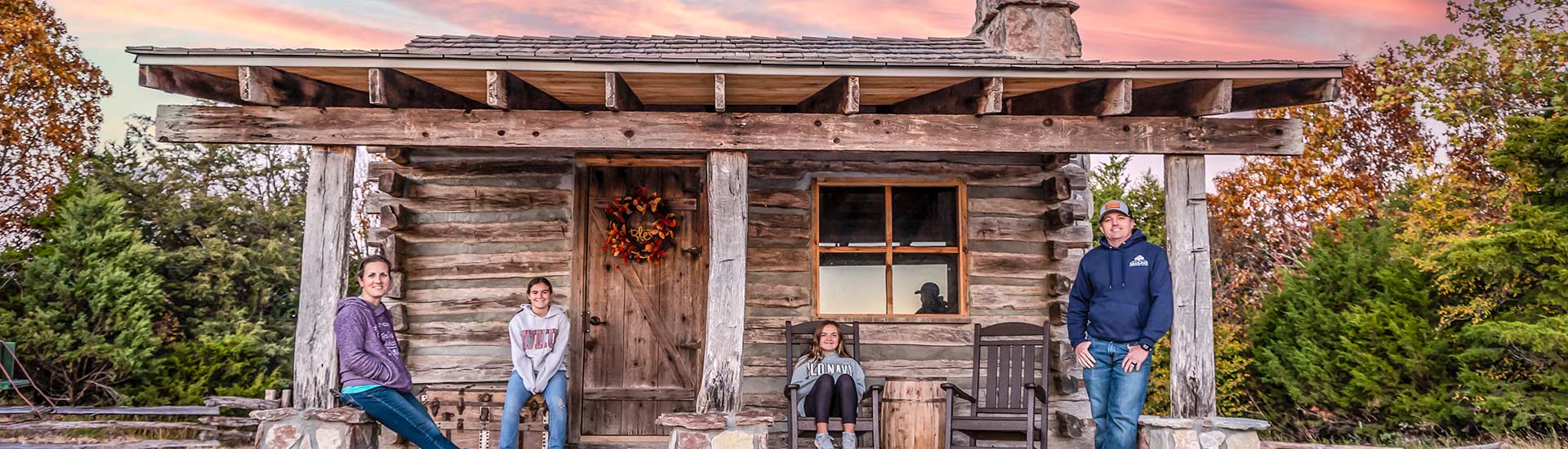 Four guests on a Pink Jeep Branson Tour pose on the porch of a historic log cabin atop Baird Mountain, lighted by a pink sunset.