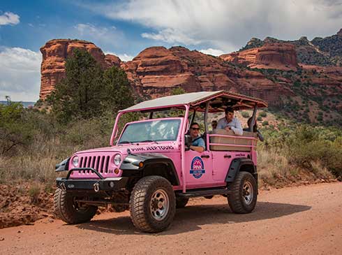Medium-shot of Pink® Jeep® with guide and guests stopped on dirt road,  Sedona's red rock formations in background