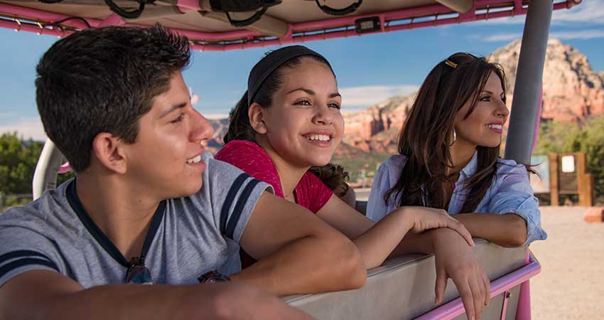 Closeup of mother and two teens looking out from the back of an open-air Pink Jeep Wrangler with Sedona's red rock landscape in the background.