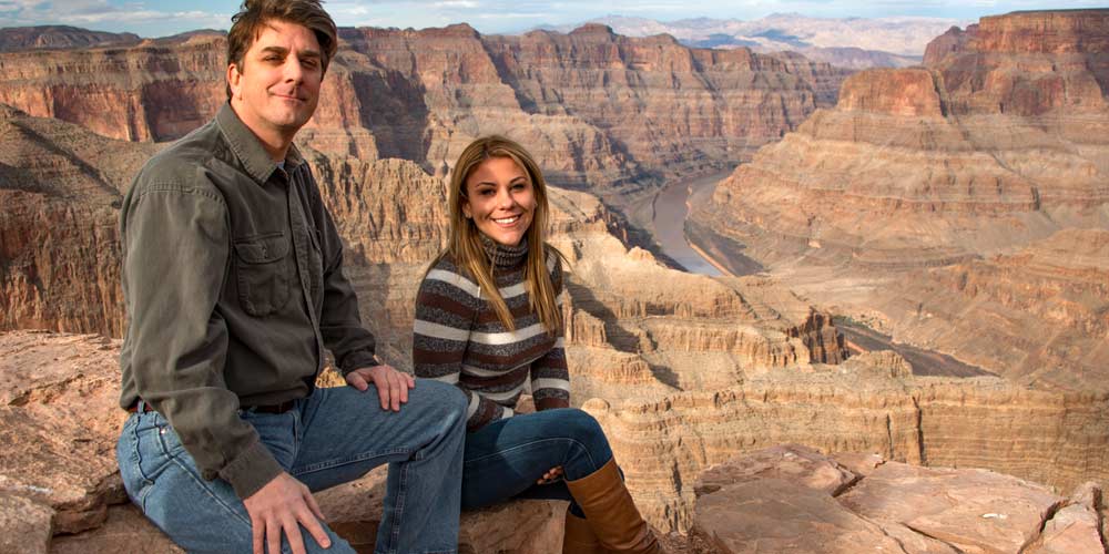 Couple looking into camera while seated on rocks overlooking the Grand Canyon's West Rim and the Colorado River below.