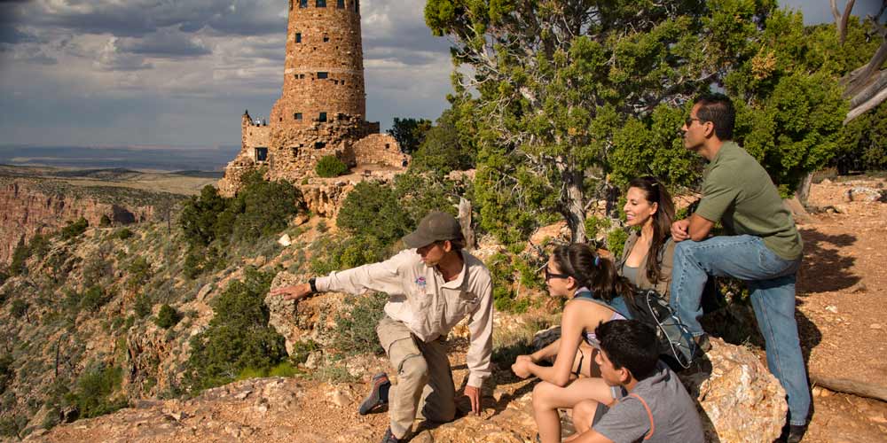 Pink Adventure tour guide kneeling beside seated guests and pointing towards Grand Canyon, with Desert View Watchtower in background.