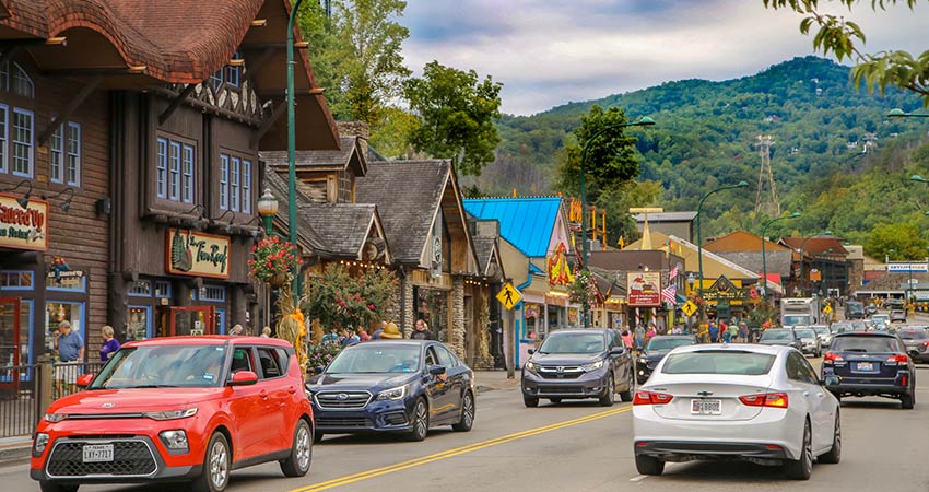 Main street in Downtown Gatlinburg during a busy summer day with blue sky and beautiful Smoky Mountains in the background.