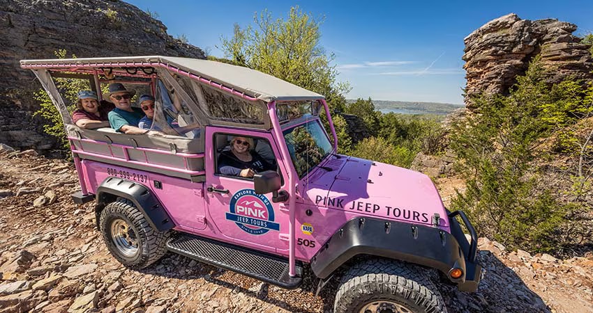 Branson tour guests smiling towards camera from back of Pink Jeep parked atop Baird Mountain with Table Rock Lake in distance.