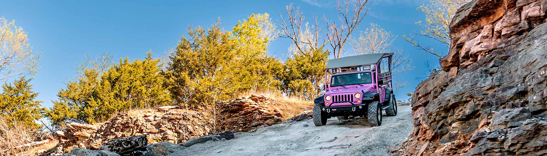 Pink Jeep Wrangler descending a steep off-road trail flanked by rock outcroppings and golden fall trees, Baird Mountain, Branson, MO.