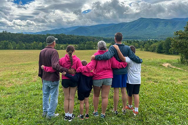 Six members of a multigenerational family stand in a Cades Cove pasture, facing the beautiful Smoky Mountains, with their backs to the camera.
