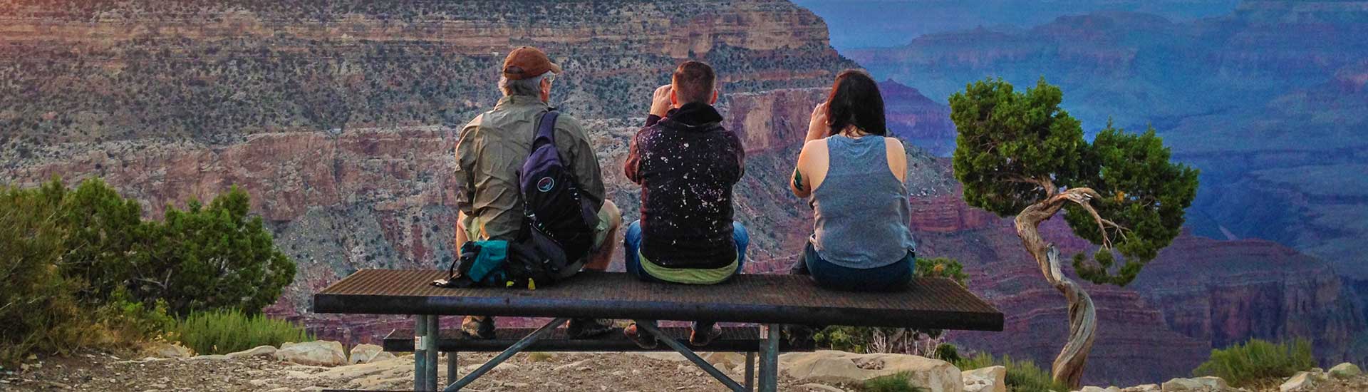 Three tourists seated atop a picnic table along the Grand Canyon South Rim, lit by the rainbow light of of an evening sunset.