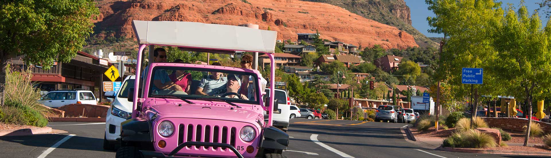 Pink Jeep Wrangler with tour guests driving towards camera with view of Uptown Sedona in the background.