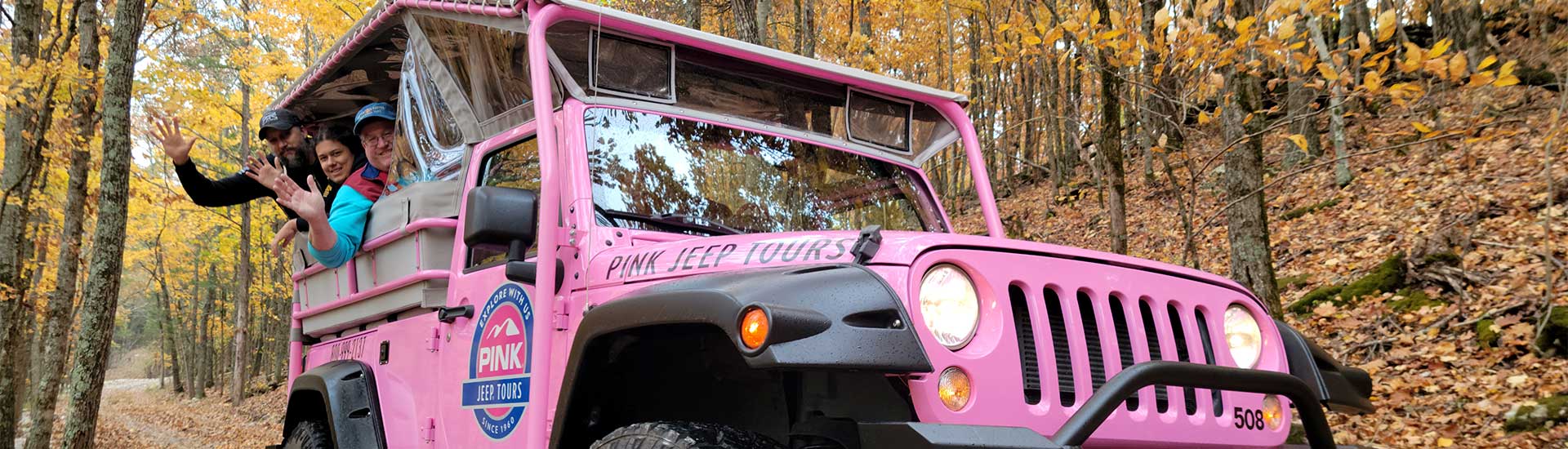 People waving from the back of a Pink Jeep Wrangler on an off-road trail flanked by autumn trees, Baird Mountain, Branson, Missouri.