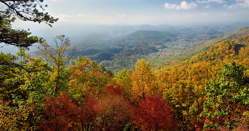 The sun rises over the mountains of Great Smoky Mountains National Park during autumn's peak, with bright red, gold and green foliage in the foreground.