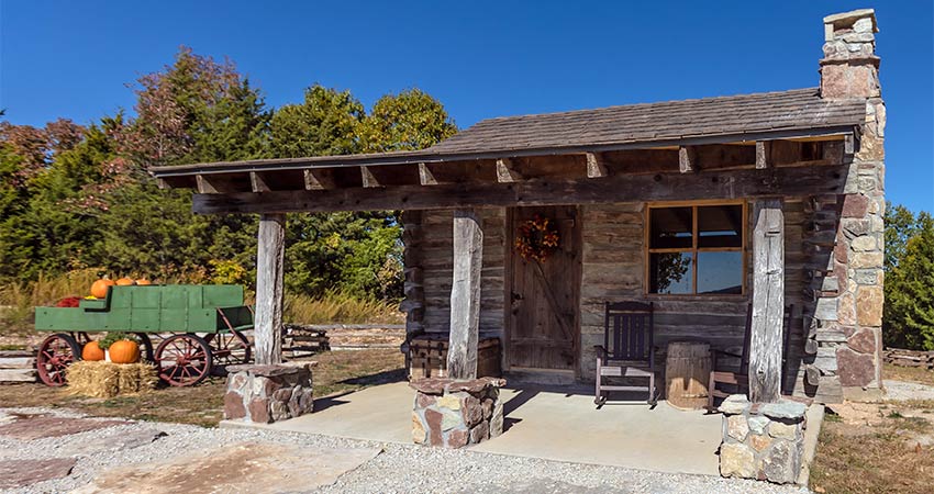 Front porch of a historic log cabin atop Baird Mountain with a pumpkin wagon parked nearby and autumn trees in background, Branson, MO.