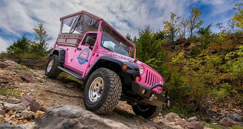 Closeup front sideview of a Pink Jeep Wrangler perched on a steep rocky trail flanked by early fall foliage and a blue sky background,