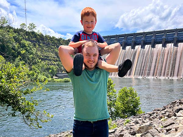 A father with a young boy on his shoulders, both smiling at the camera, standing near the spillway of Table Rock Dam in Branson, Missouri.