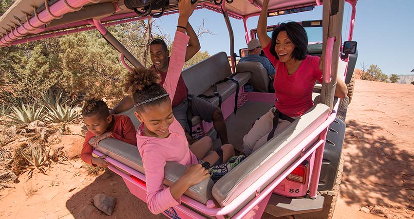 Closeup of a family of four seated in the back of a Pink Jeep Wrangler and holding the rails, while navigating a steep incline on the Broken Arrow Trail.