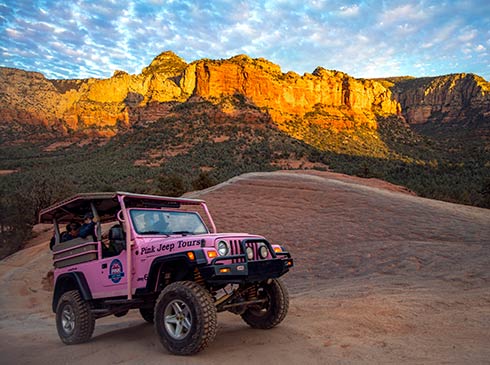 Pink Jeep Wrangler with guests driving across Sedona's slickrock with surrounding red rock formations glowing during an evening sunset.