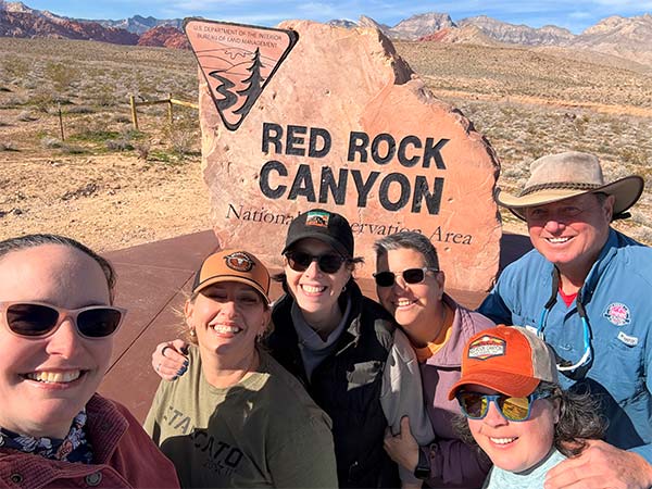 Pink Jeep Tours Las Vegas adventure guide poses with a group of women in front of the Red Rock Canyon National Conservation Area stone cut sign.