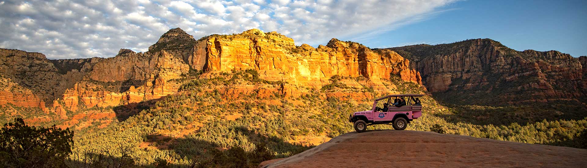 Panoramic view Sedona's red rock formations glowing in golden hues just before sunset with a Pink Jeep Wrangler perched atop slickrock in the foreground.