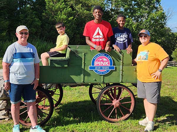 Two men and three young boys pose by a green, vintage wooden wagon with the Branson Pink Jeep Tours logo sign centered on its side, Branson, MO.